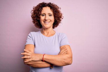 Orta yaşlı güzel kıvırcık saçlı kadın izole edilmiş pembe arka planda tişört giyip kollarını kavuşturup kameraya bakarken gülümsüyor. Pozitif kişi.