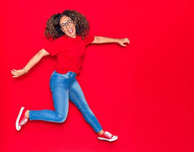 Orta yaşlı, güzel İspanyol bir kadın günlük kıyafetler ve mutlu bir gülümseme takınıyor. İzole edilmiş kırmızı arka planda yüzünde gülümsemeyle zıplıyor