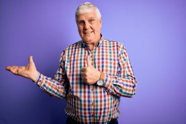 Kıdemli yakışıklı, beyaz gömlekli, soyutlanmış mor arka planda avuçlarını gösteren ve baş parmaklarıyla hareket eden mutlu ve neşeli gülümseyen bir adam.