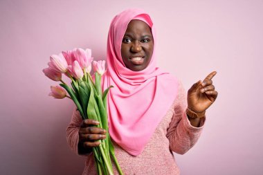 Genç Afro-Amerikan artı iri kadın elinde pembe laleler buketi tutan Müslüman başörtüsü taşıyor endişeli ve gergin bir kenara işaret eden işaret parmağı, endişeli ve şaşırmış yüz ifadesi