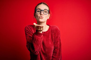 Genç, güzel, kızıl saçlı bir kadın izole edilmiş kırmızı arka plan üzerine gündelik süveter giyip kameraya bakıyor ve havada bir öpücük savuruyor. Güzel ve seksi. Aşk ifadesi.