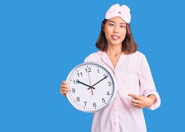 Uyku maskesi ve pijama giyen genç ve güzel Çinli kız elinde kocaman bir saat tutarak mutlu bir şekilde elini ve parmağını işaret ediyor. 