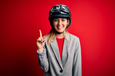 Kırmızı arka planda motosiklet kaskı takan güzel sarışın motosikletli kadın kendini beğenmiş ve mutlu bir şekilde gülümserken bir numarayı işaret ediyor..