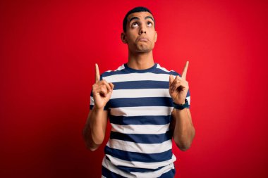 Yakışıklı Afro-Amerikan erkeği kırmızı arkaplan üzerinde sıradan çizgili tişört giyiyor üzgün ve üzgün görünüyor, parmaklarıyla yönü gösteriyor, mutsuz ve depresif..