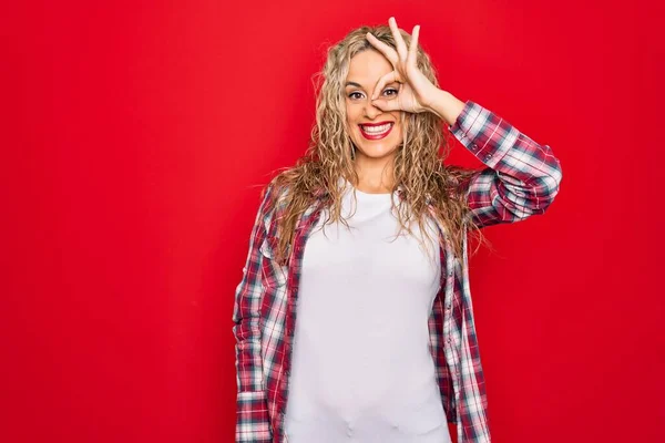 Young beautiful blonde woman wearing casual shirt standing over isolated red background doing ok gesture with hand smiling, eye looking through fingers with happy face.