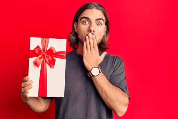 Young handsome man holding gift covering mouth with hand, shocked and afraid for mistake. surprised expression