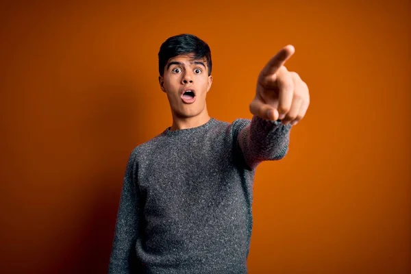 Young handsome man wearing casual sweater standing over isolated orange background Pointing with finger surprised ahead, open mouth amazed expression, something on the front