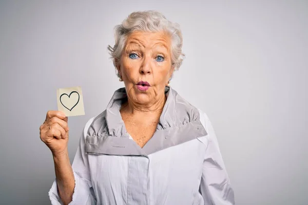 Senior beautiful grey-haired woman holding reminder paper heart over white background scared in shock with a surprise face, afraid and excited with fear expression