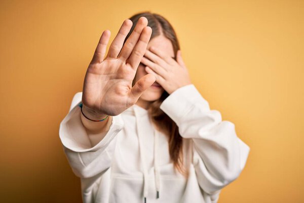 Young beautiful redhead sporty woman wearing sweatshirt over isolated yellow background covering eyes with hands and doing stop gesture with sad and fear expression. Embarrassed and negative concept.