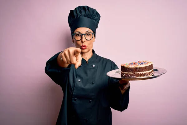 Middle age baker woman wearing cooker uniform and hat holding tray with delicious cake pointing with finger to the camera and to you, hand sign, positive and confident gesture from the front