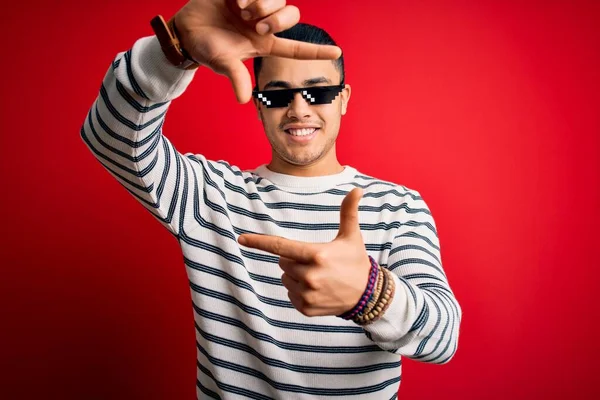 年轻的巴西男人戴着滑稽暴徒的生活太阳镜 在孤立的红色背景上微笑着 用双手和手指头做着快乐的框架 创意和摄影概念 — 图库照片