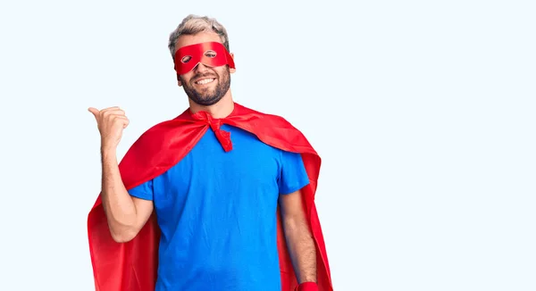 Young blond man wearing super hero custome smiling with happy face looking and pointing to the side with thumb up.