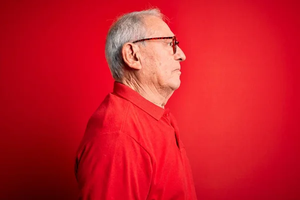 Grey haired senior man wearing glasses and casual t-shirt over red background looking to side, relax profile pose with natural face with confident smile.