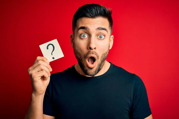 Young handsome man holding paper with question mark symbol over red background scared in shock with a surprise face, afraid and excited with fear expression