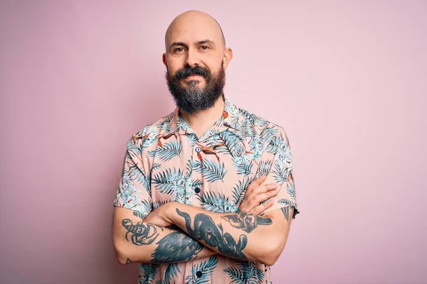 Sakallı, dövmeli, pembe arka planda sıradan bir çiçek gömleği giyen, kollarını kavuşturup kameraya bakan yakışıklı kel adam. Pozitif kişi.