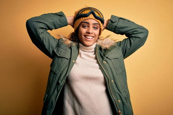 Genç Afrikalı Afro-Amerikan kayakçı kız kar sporu kıyafetleri ve kayak gözlükleri takıyor rahatlıyor ve esniyor, kolları ve elleri başının ve boynunun arkasında mutlu bir şekilde gülümsüyor.