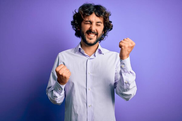 Young handsome business man with beard wearing shirt standing over purple background celebrating surprised and amazed for success with arms raised and eyes closed. Winner concept.