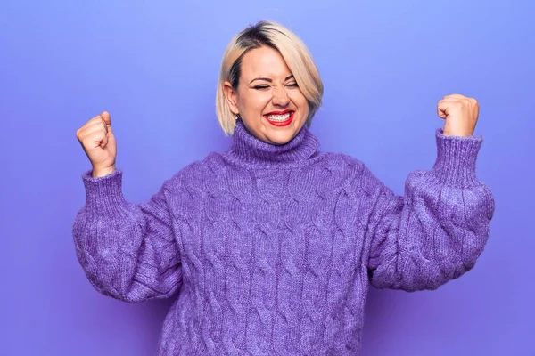 Güzel sarışın ve boyunda bir kadın mor arka plan üzerine balıkçı yaka kazak giyiyor. Çok mutlu ve heyecanlı. Kolları havada, gülümseyen ve başarı için çığlık atan kazanan bir jest yapıyor. Kutlama konsepti.