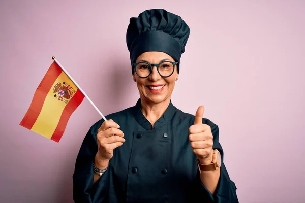 中年爱国女厨师身穿炊事服 头戴西班牙色彩旗 喜形于色 笑着做手势 用手指竖起大拇指 是极好的标志 — 图库照片