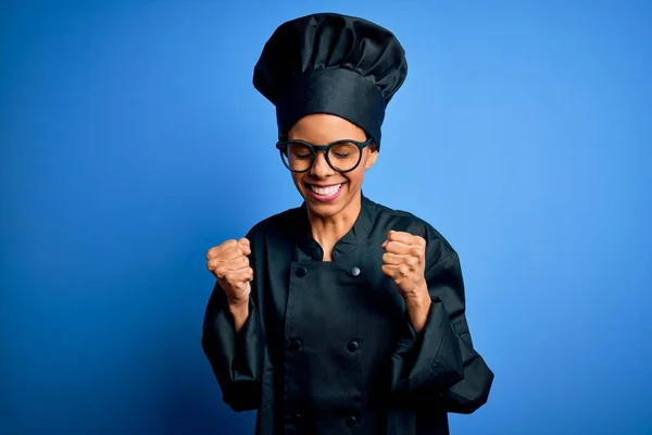 年轻的非洲女厨师身穿炊具制服 头戴蓝色背景的帽子 兴奋地期待着胜利 举起双臂 闭上双眼 微笑着庆祝胜利 赢的概念 — 图库照片