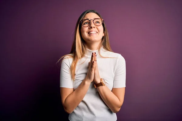 紫色の背景にカジュアルなTシャツを着た青い目の若い美しいブロンドの女性が物乞いをし 手で祈るとともに 顔に希望の表情を非常に感情的で心配しています 物乞い — ストック写真