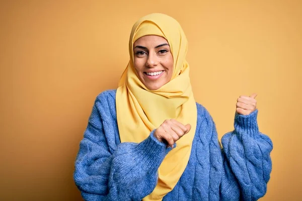 Young beautiful brunette muslim woman wearing arab hijab over isolated yellow background Pointing to the back behind with hand and thumbs up, smiling confident