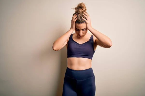 Young beautiful blonde sportswoman doing sport wearing sportswear over white background suffering from headache desperate and stressed because pain and migraine. Hands on head.