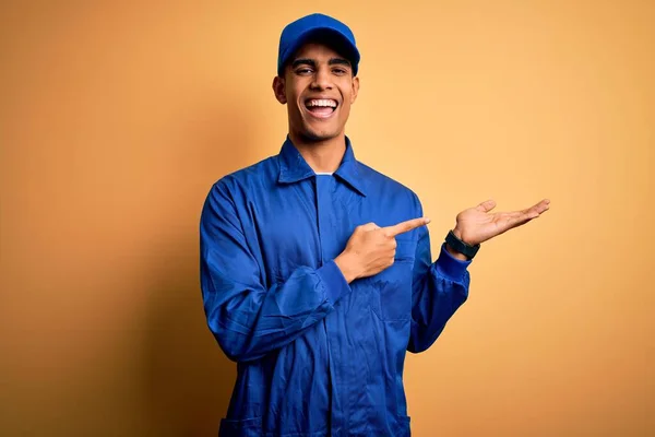身穿蓝色制服 头戴黄色背景帽的年轻的非洲机械师惊奇地对着摄像机微笑 同时用手和手指指点着镜头 — 图库照片