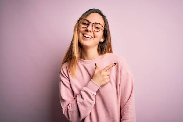 Beautiful blonde woman with blue eyes wearing sweater and glasses over pink background cheerful with a smile of face pointing with hand and finger up to the side with happy and natural expression on face