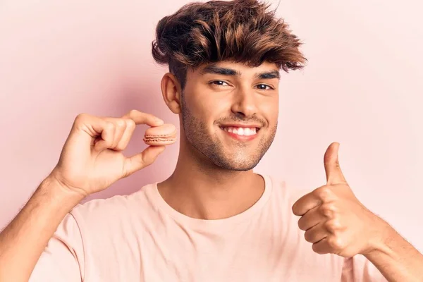 Joven Hombre Hispano Sosteniendo Macaron Sonriendo Feliz Positivo Pulgar Hacia — Foto de Stock