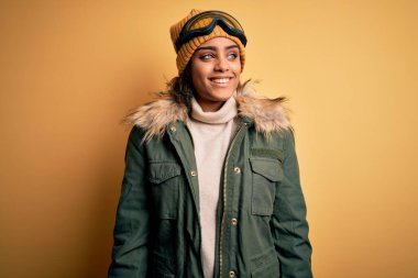 Genç Afro-Amerikan kayakçı kız kar sporu giyer ve kar gözlüğü takar yüzünde gülümseme, doğal bir ifade ile yan yana bakar. Kendinden emin gülen.