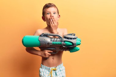 Mayo giyen sevimli sarışın çocuk elinde su tabancasıyla ağzını eliyle kapatıyor, şok geçiriyor ve hata yapmaktan korkuyor. şaşırmış ifade 