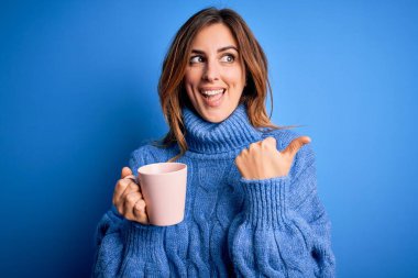 Genç, güzel, esmer bir kadın izole edilmiş mavi arka planda bir fincan kahve içiyor ve gülümseyen bir gülümsemeyle baş parmağıyla işaret ediyor.