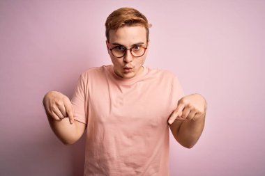 Genç, kızıl saçlı, günlük tişört giyen, pembe arka plan üzerinde duran, parmaklarını göstererek reklamını yapan, şaşırmış yüzünü ve ağzını açan genç bir adam.