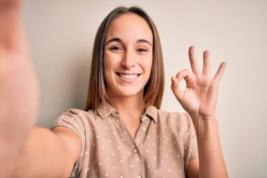 Yazlık tişört giyen genç ve güzel bir kadın beyaz arka planda kamerayla selfie çekiyor. Tamam işareti yapıyor, mükemmel bir sembol.