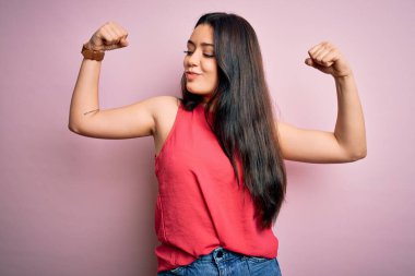 Genç esmer kadın pembe izole edilmiş arka plan üzerine gündelik bir tişört giyiyor ve gururla gülümseyen kol kaslarını gösteriyor. Fitness konsepti.