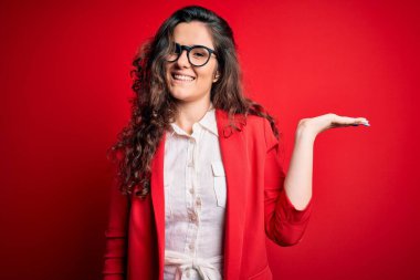 Kıvırcık saçlı, ceketli ve kırmızı arka planda gözlüklü neşeli bir şekilde gülümseyen ve avuç içi kameraya bakan güzel bir kadın..
