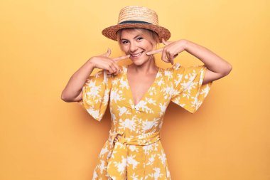 Güzel sarışın kadın tatilde sarı arka planda giyinmiş ve şapkasını takmış gülümseyen ve parmaklarıyla ve ağzıyla işaret eden. Diş sağlığı konsepti.