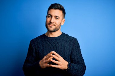 İzole edilmiş mavi arka plan üzerinde duran sıradan bir süveter giyen genç adam ellerini birleştirip gülümseyerek gevşemiş ve neşeli olmasını diliyordu. Başarı ve iyimser