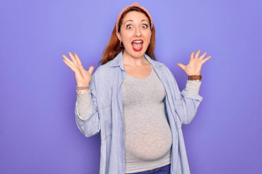 Genç, güzel, kızıl saçlı hamile bir kadın mor arka planda bebek bekliyor. Çılgınlığı kutluyor ve başarıyı kollarını kaldırıp heyecanla haykırarak kutluyor. Kazanan konsept