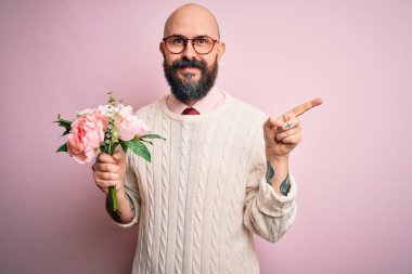 Yakışıklı, kel ve sakallı bir adam pembe arka planda güzel bir buket tutuyor.