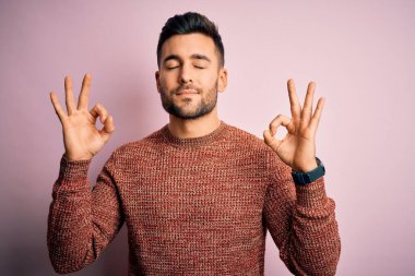 İzole edilmiş pembe arka plan üzerinde duran sıradan süveter giyen genç yakışıklı adam rahatla ve parmaklarıyla meditasyon hareketi yaparken gözleri kapalı gülümse. Yoga konsepti.