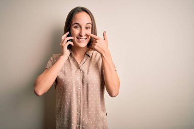 Genç ve güzel bir kadın beyaz arka planda akıllı telefondan konuşuyor. El ve burunla işaret ediyor. Neşeli gülümsüyor. Güzellik konsepti