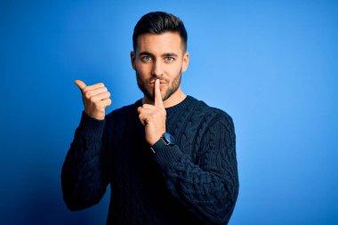 İzole edilmiş mavi arka plan üzerinde sıradan bir süveter giyen genç ve yakışıklı bir adam ellerini yana uzatarak parmaklarını dudaklarına dayayarak sessiz olmak istiyor. Sessizlik ve gizli konsept.
