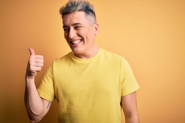 Sarı izole edilmiş arka planda sarı tişört giyen genç, yakışıklı ve modern bir adam gülümseyen bir yüz ifadesiyle başparmağıyla yan tarafa bakıyor..