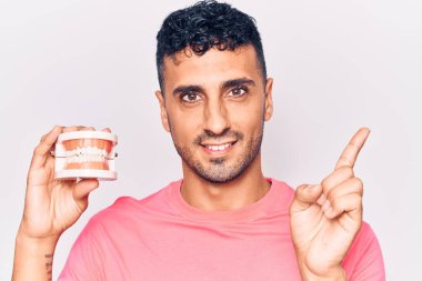 Takma diş tutan genç İspanyol adam gülümsüyor mutlu bir şekilde elini ve parmağını yana doğru gösteriyor. 