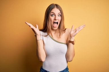 Genç, güzel, kızıl saçlı bir kadın izole edilmiş sarı arka plan üzerine gündelik tişört giyip çılgınlığı kutluyor ve başarıya hayret ediyor. Kolları havada ve gözleri açık, heyecanlı bir şekilde çığlık atıyor. Kazanan konsept