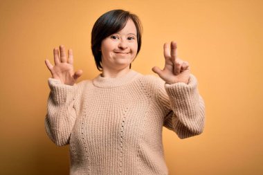 Sarı arka planda rahat süveter giyen genç Down sendromlu kadın sekiz numaralı parmaklarıyla gülümseyerek kendinden emin ve mutlu bir şekilde onu işaret ediyor..