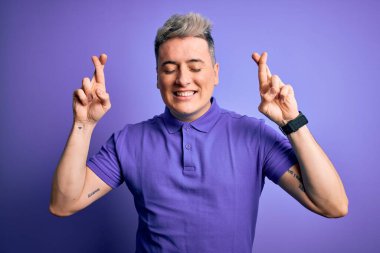 Genç, yakışıklı, modern bir adam izole edilmiş arka plan jestlerinin üzerine gündelik mor tişört giyip umut ve gözler kapalı gülümseyerek el sallıyor. Şans ve batıl inanç..