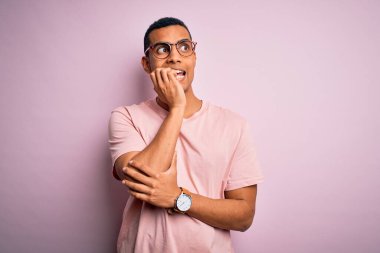 Yakışıklı Afro-Amerikalı adam pembe arka planda günlük tişört ve gözlük takıyor. Ağzı tırnak ısıran elleri yüzünden gergin ve stresli görünüyor. Endişe sorunu.